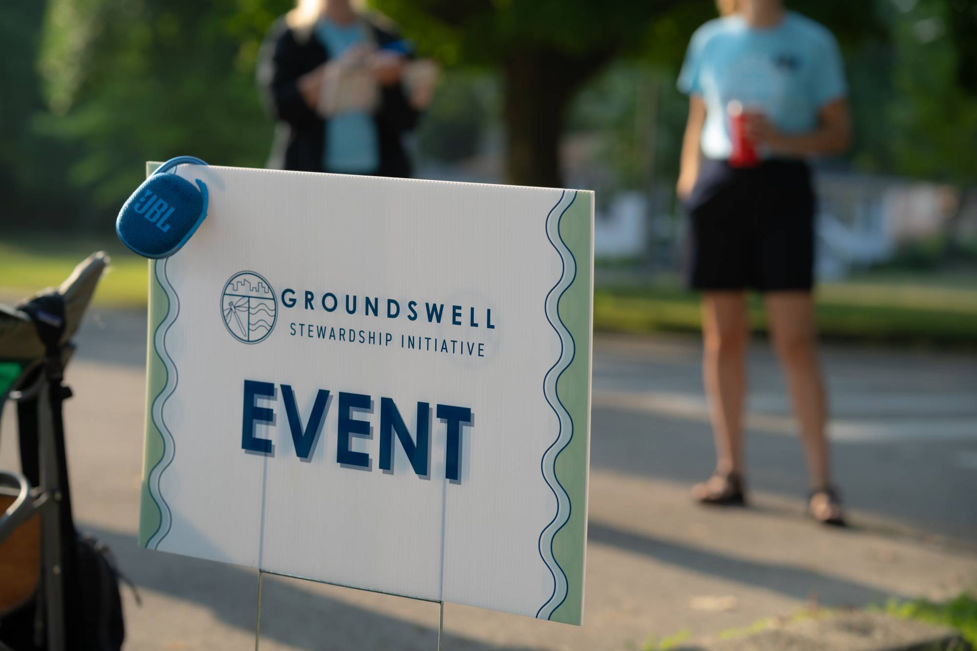 close up on sign that reads "event" with the groundswell logo. People standing in the sunshine in the background.